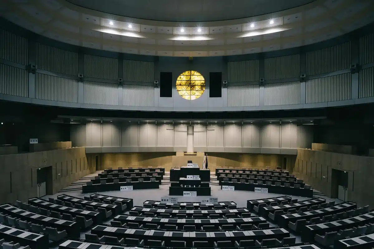 Cámara parlamentaria con legisladores debatiendo en asientos, arquitectura histórica de fondo.
